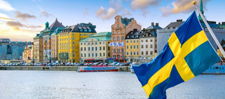 The flag of Sweden with a city in the background. The site is on a harbour.