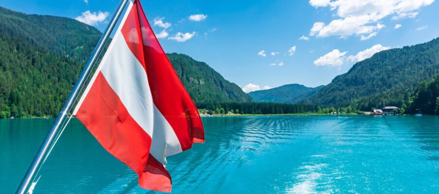The flag of Austria with a blue sky, a lake and mountains in the background.
